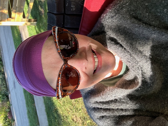 Dr. Lange smiles while sitting outside. She is wearing a grey shawl, a purple bandana on her head, and funky brown cat eye sunglasses.