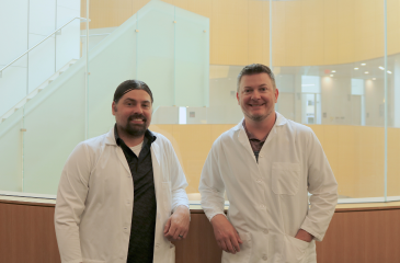 Branden Moriarity (left) and Beau Webber (right) pose in front of the wood and glass wall at the CCRB building on the UMN Twin Cities campus. They are both wearing white coats and smiling as they lean back on the wall behind them.