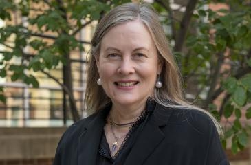 Dr. Carol Lange poses for a headshot outside. Behind her, there are trees with green leaves on them. Her hair is long and sandy blonde. She is wearing pearl earrings and a black jacket top.